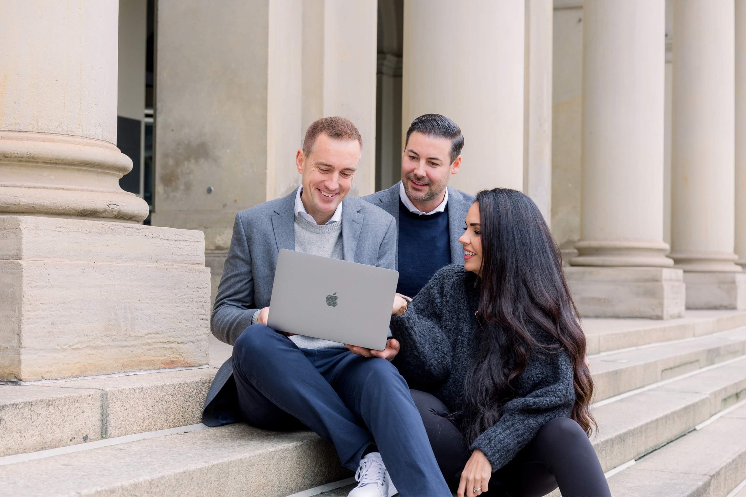 Three businesspeople sitting on steps outside, looking at a laptop and smiling.