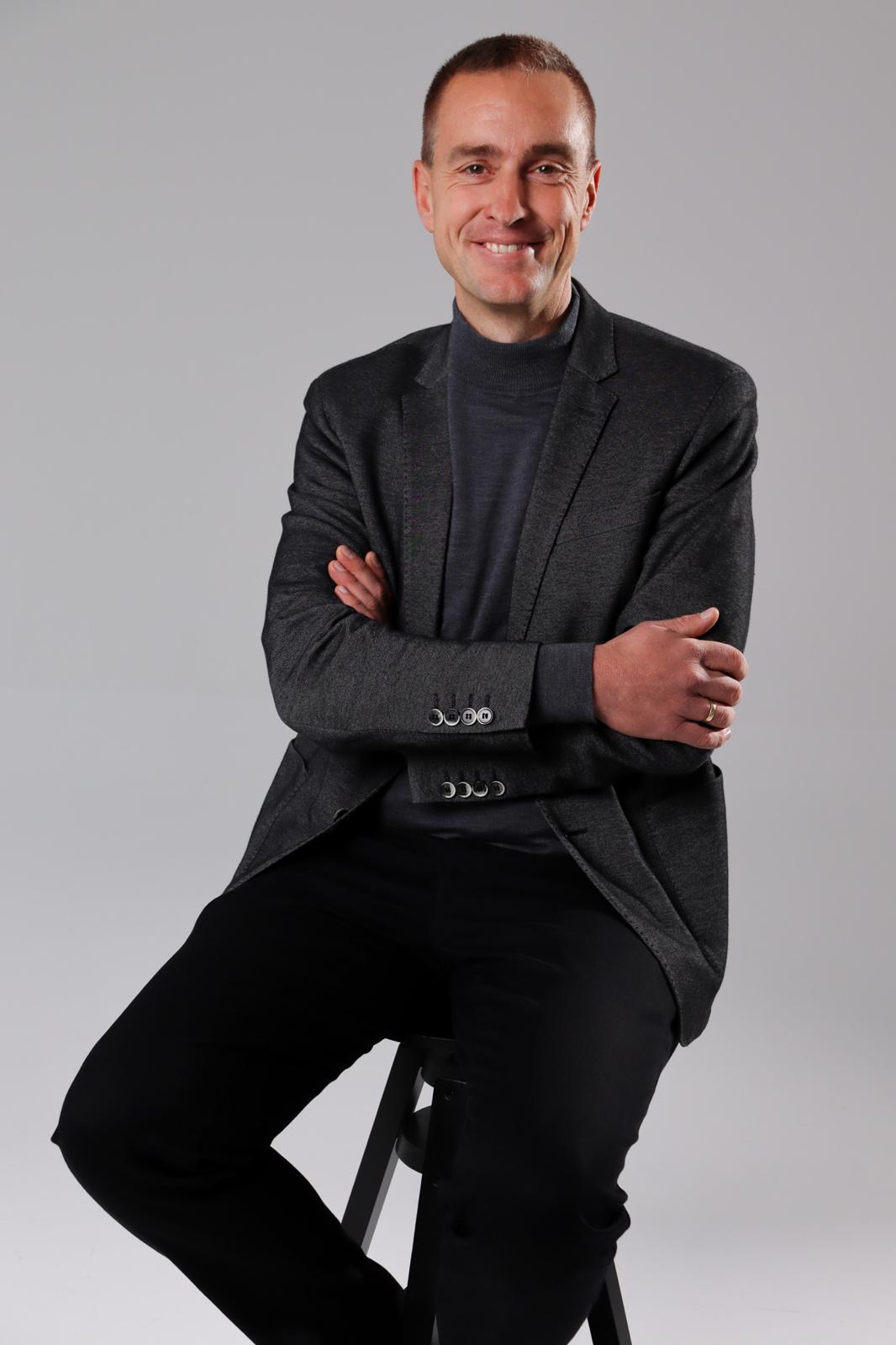 Man in dark blazer and turtleneck sitting on a stool with arms crossed, smiling at camera.