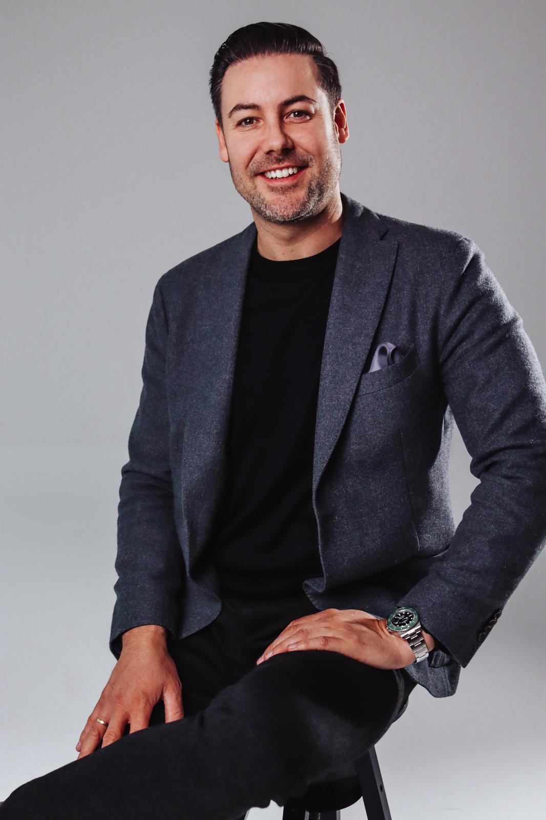 Smiling man sitting on stool wearing a grey blazer, black shirt, and watch against gray background.