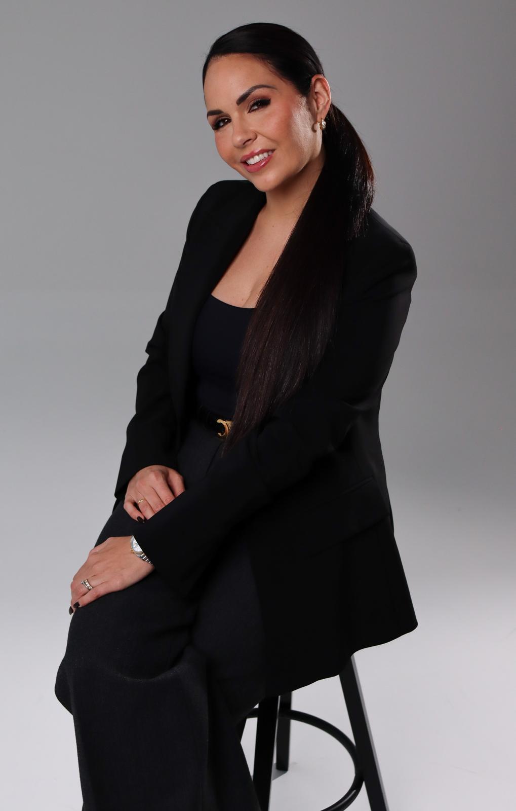 Woman with long dark hair in a black blazer sitting on a black stool against a gray background.