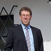Man wearing glasses, a dark suit, light blue shirt, and striped tie standing outside near a window.