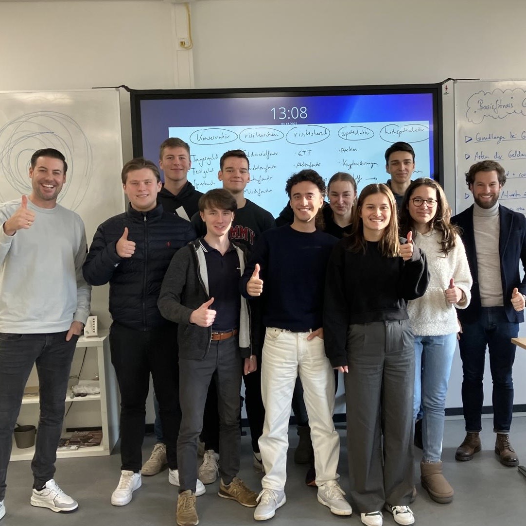 Group of ten young adults smiling indoors, giving thumbs up in front of a whiteboard and screen.