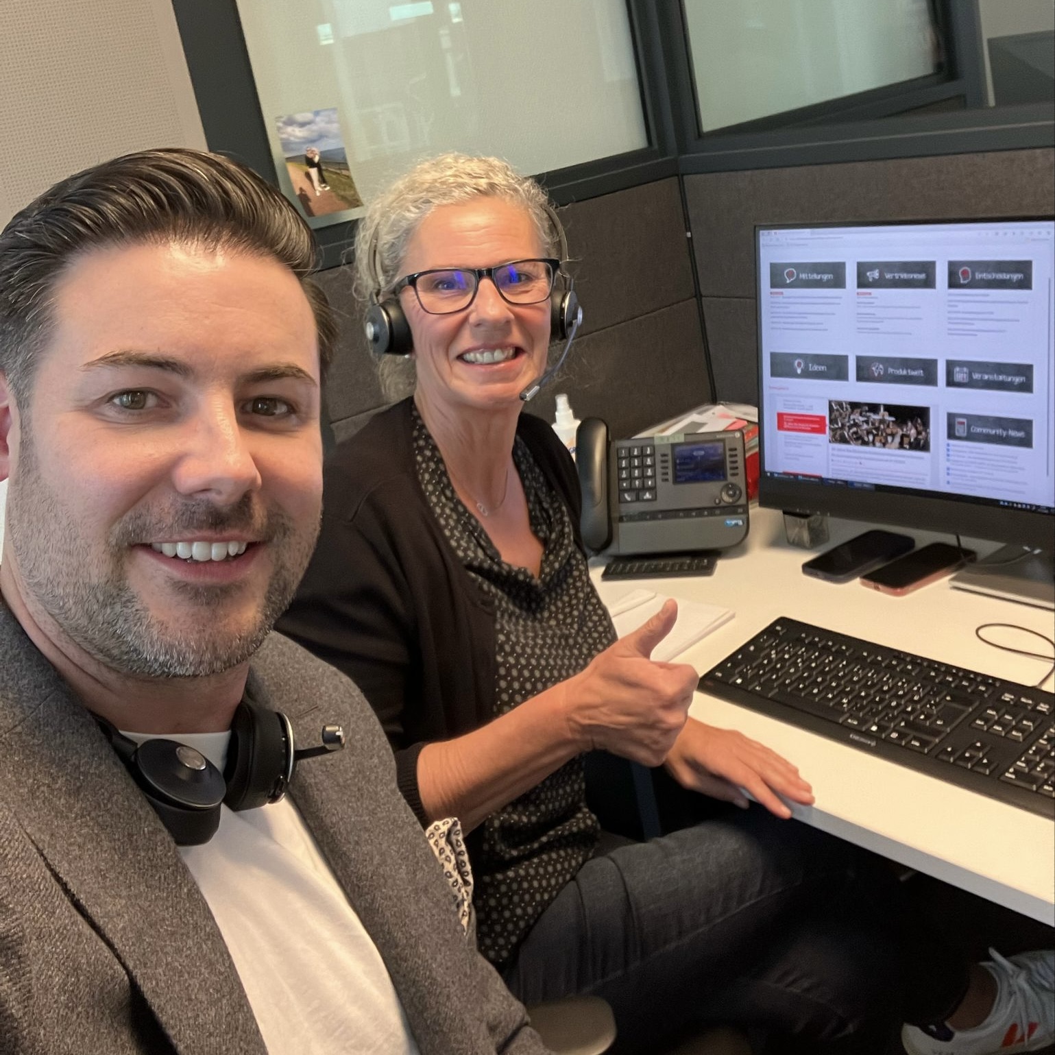 Man and woman with headsets smiling at office desk with computer and phone, woman giving thumbs up.