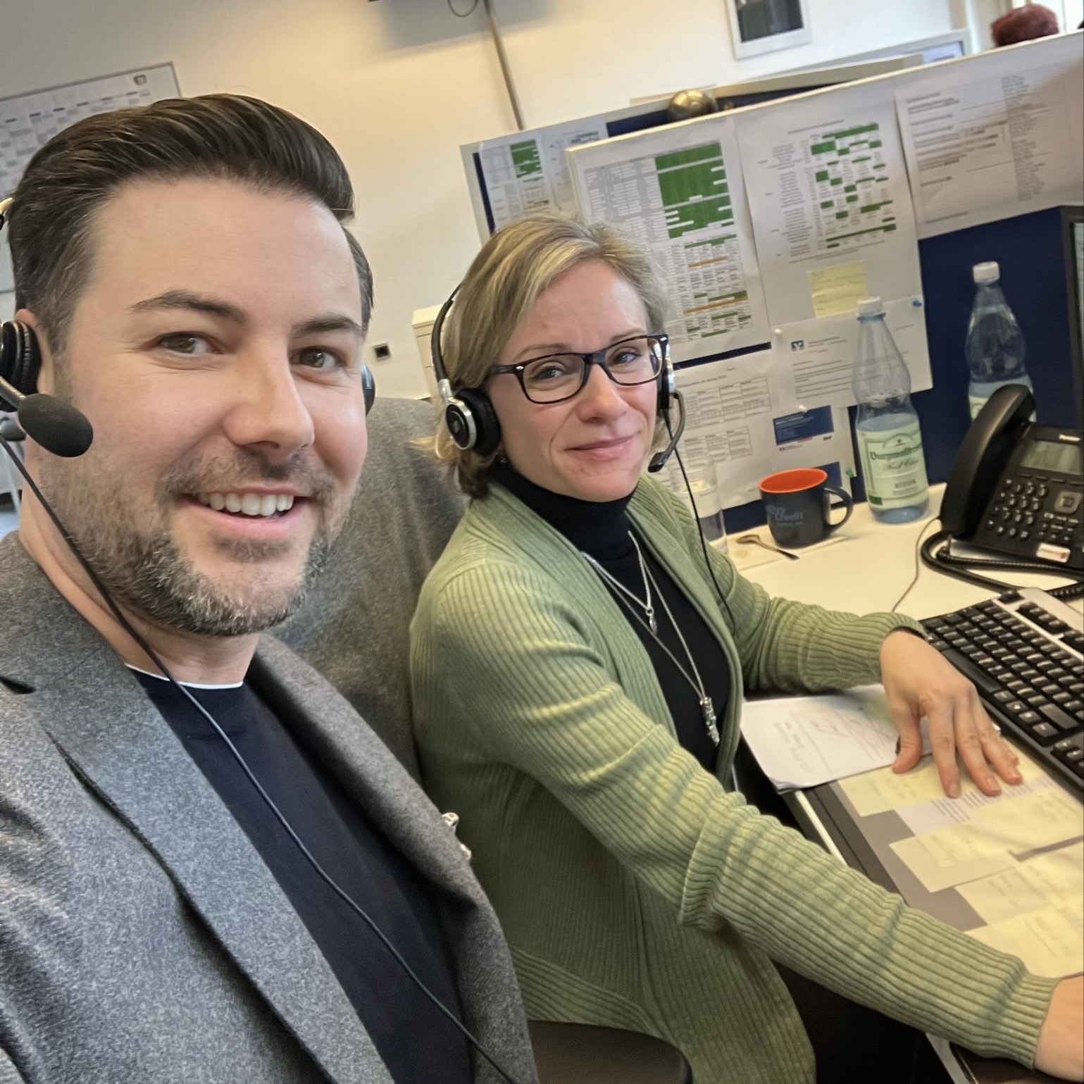 Man and woman wearing headsets sitting at an office desk with computer and papers.