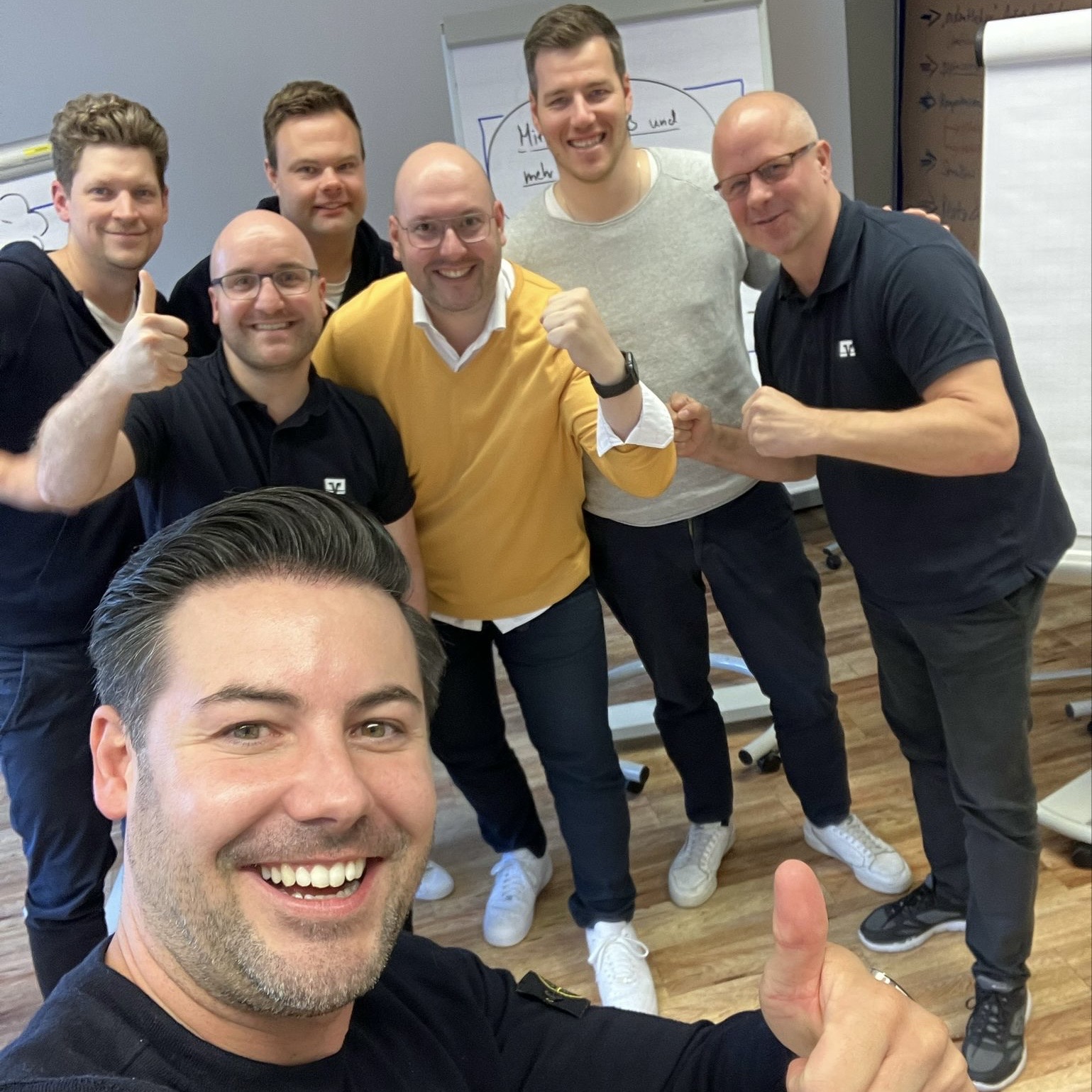 Seven men smiling and posing in a room, some with thumbs up or fist gestures, standing near flip charts.
