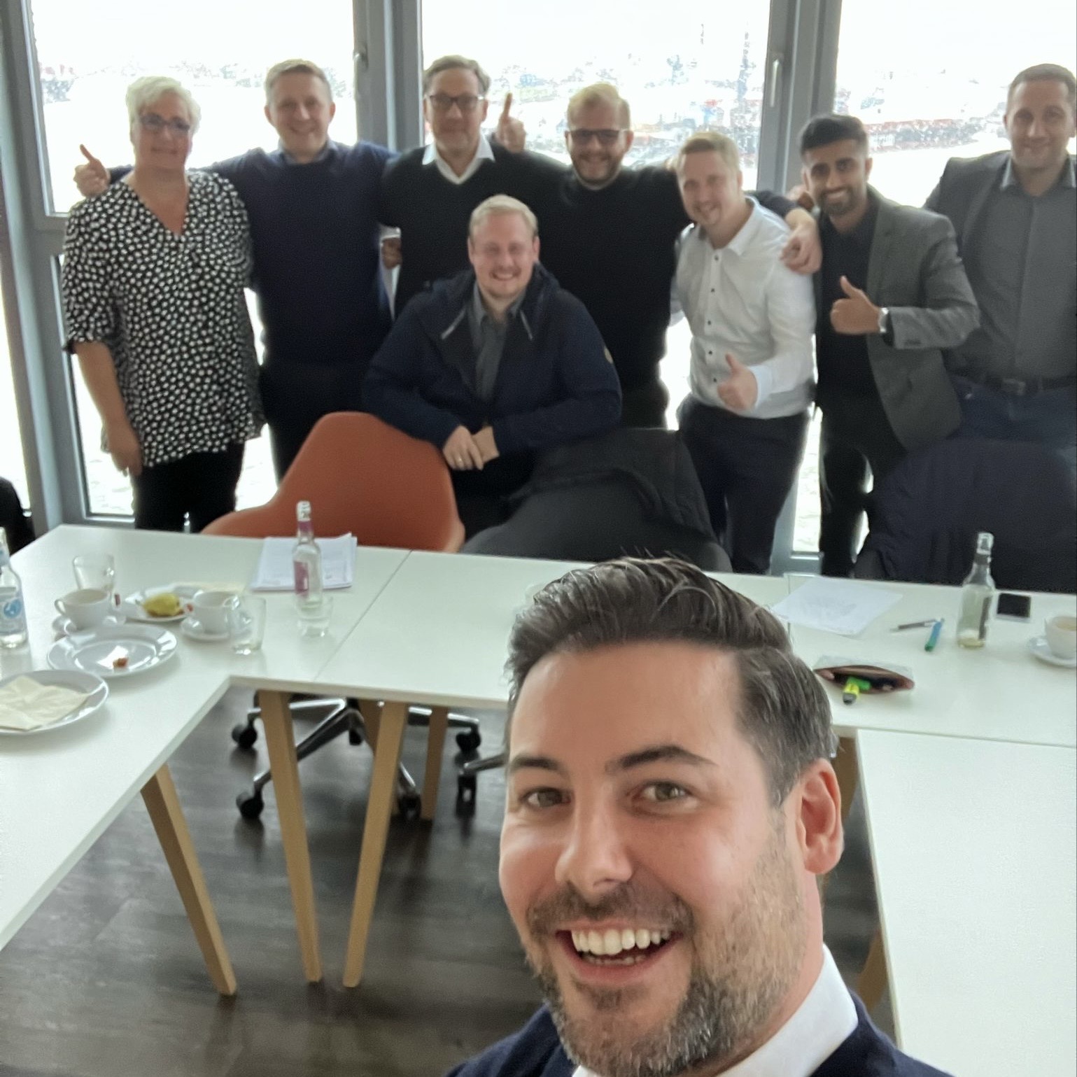 Group of nine smiling people posing together indoors with a man taking a front selfie.