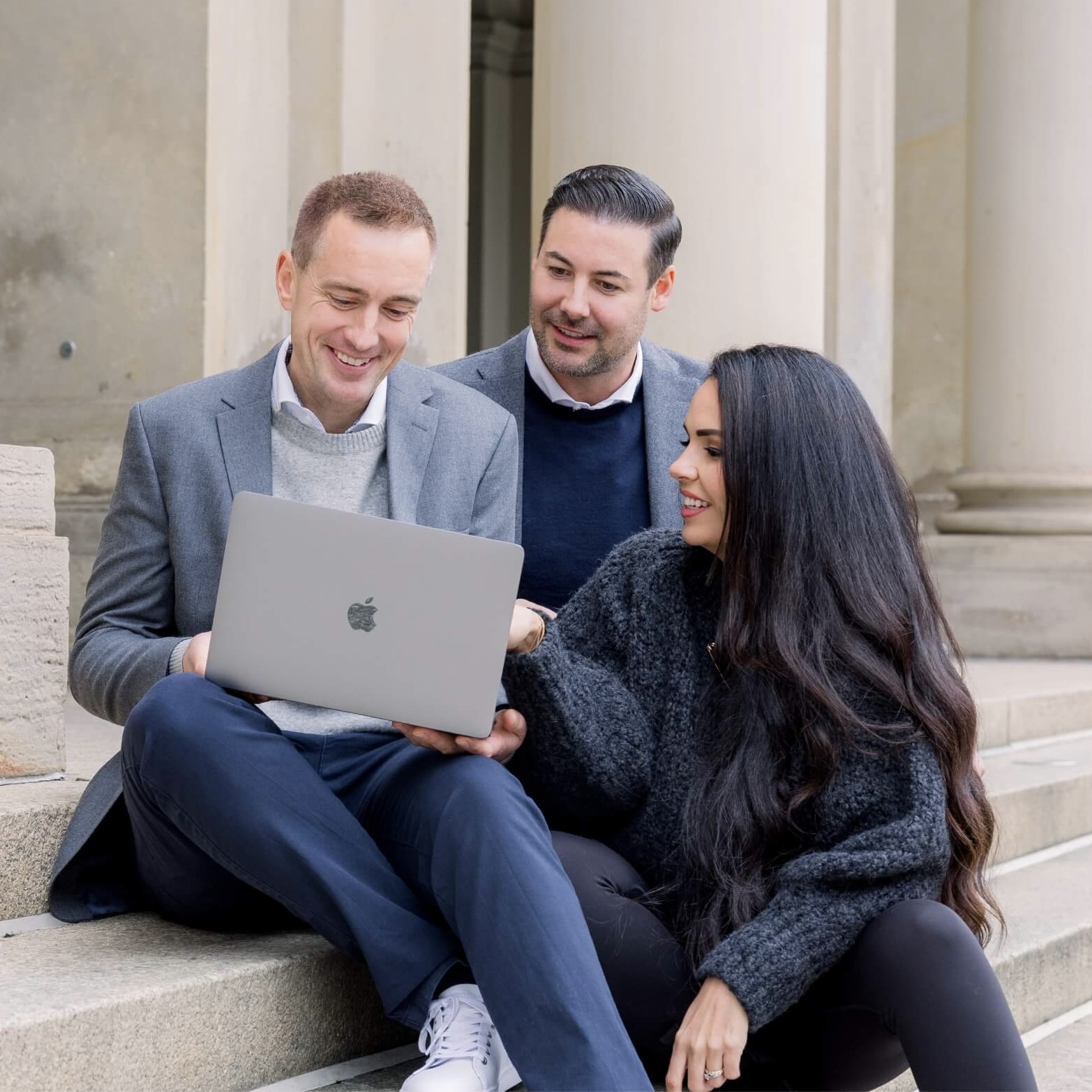 Two men and one woman sitting on stone steps, smiling, looking at a laptop held by a man.