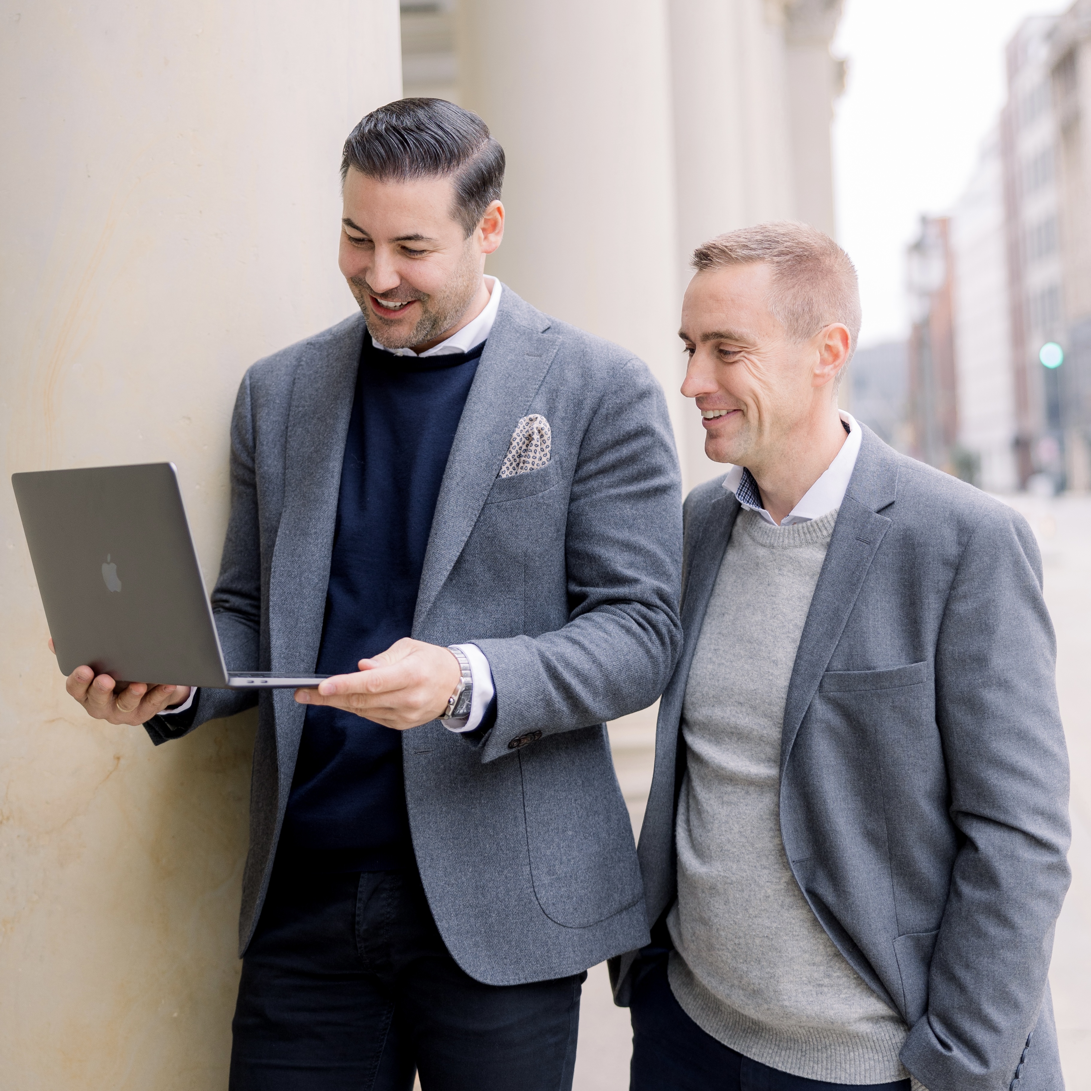 Two businessmen in gray blazers smiling while looking at a laptop outside near pillars.