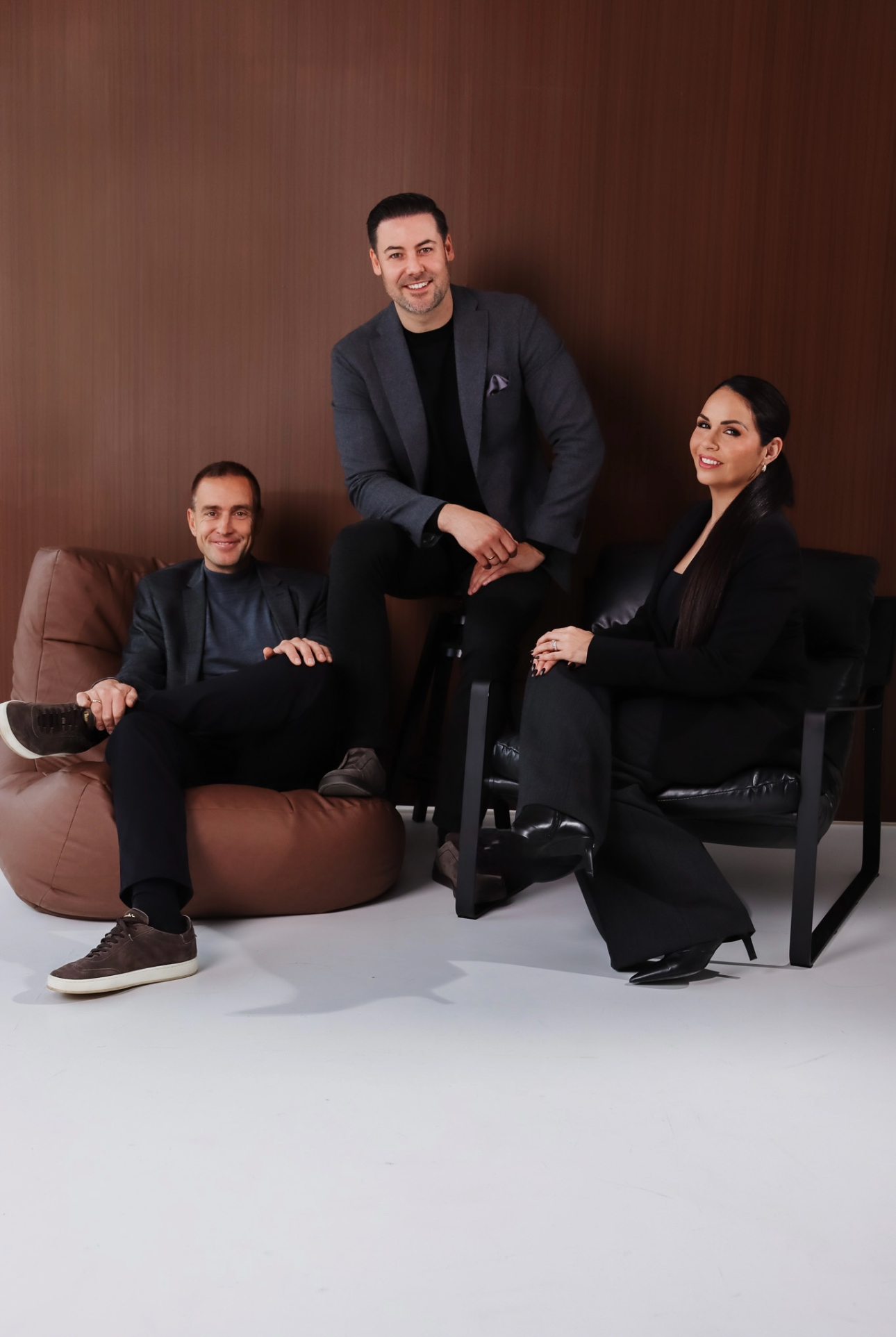 Two men and a woman dressed in dark clothing posing seated on brown beanbag, black chair, and stool.