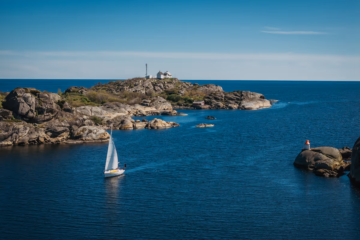 Sailboat on calm blue water near rocky island with houses under blue sky.