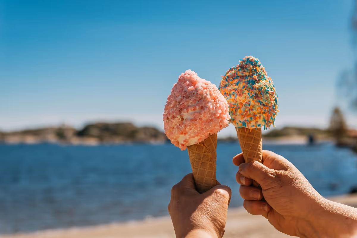 Two hands holding ice cream cones with pink and multicolored sprinkles by the water under a blue sky.