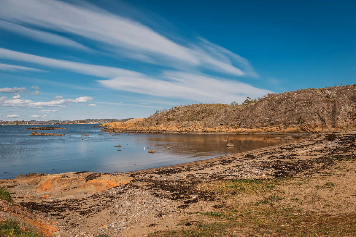 Calm coastal scene with rocky shore, patches of grass, and blue sky with streaked clouds.