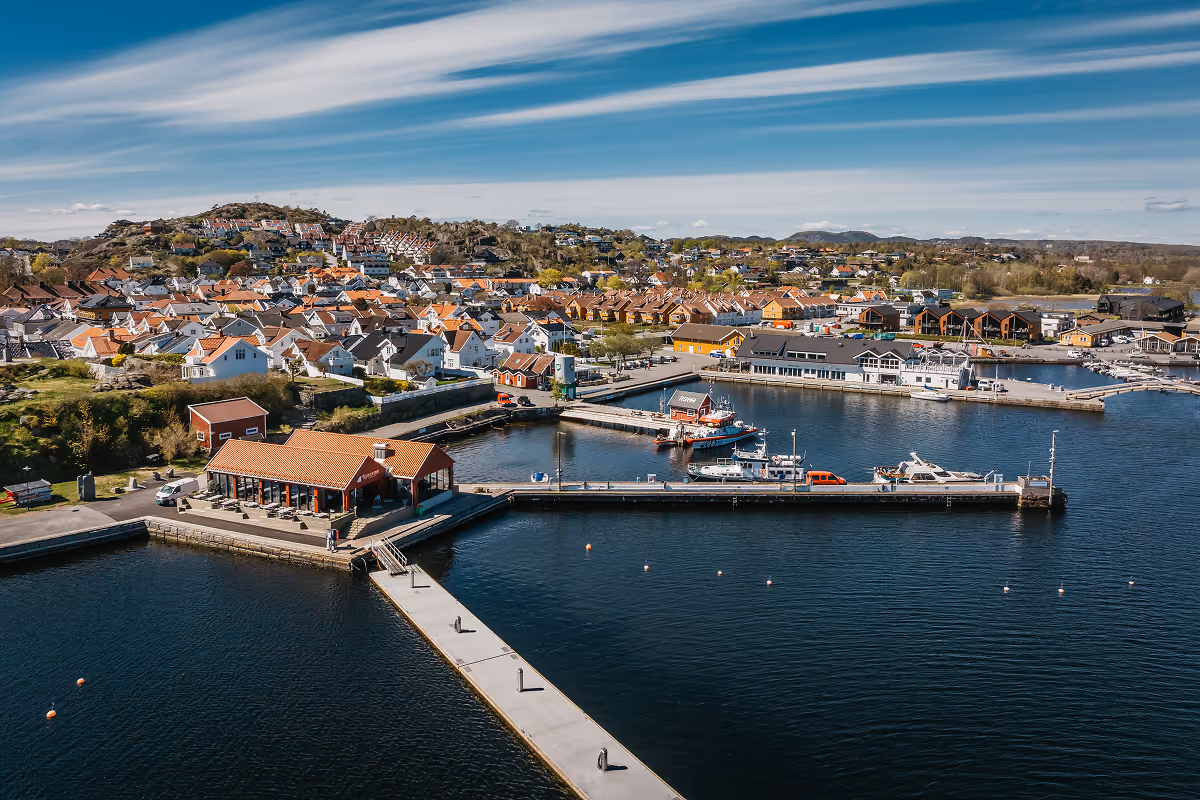 Aerial view of a harbor with docks, boats, and residential houses on rolling hills under a blue sky.