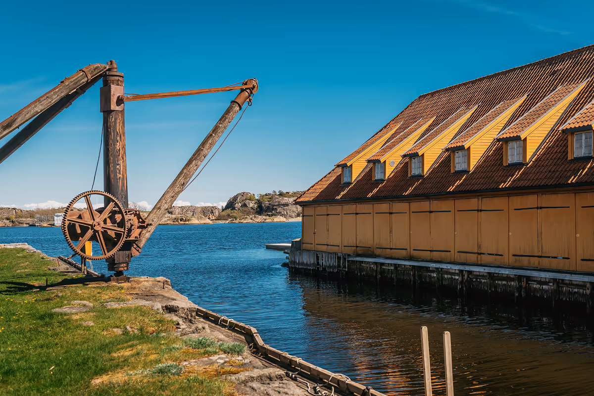 Old wooden crane by water next to yellow building with multiple windows under blue sky.