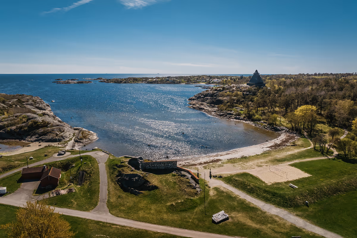 Coastal bay with rocky shores, sandy beach, green grass, trees, and a pyramid-shaped building on the right.