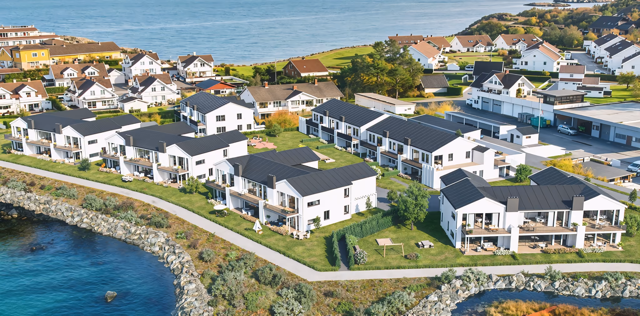 Aerial view of coastal neighborhood with modern white houses, green lawns, and rocky shore.