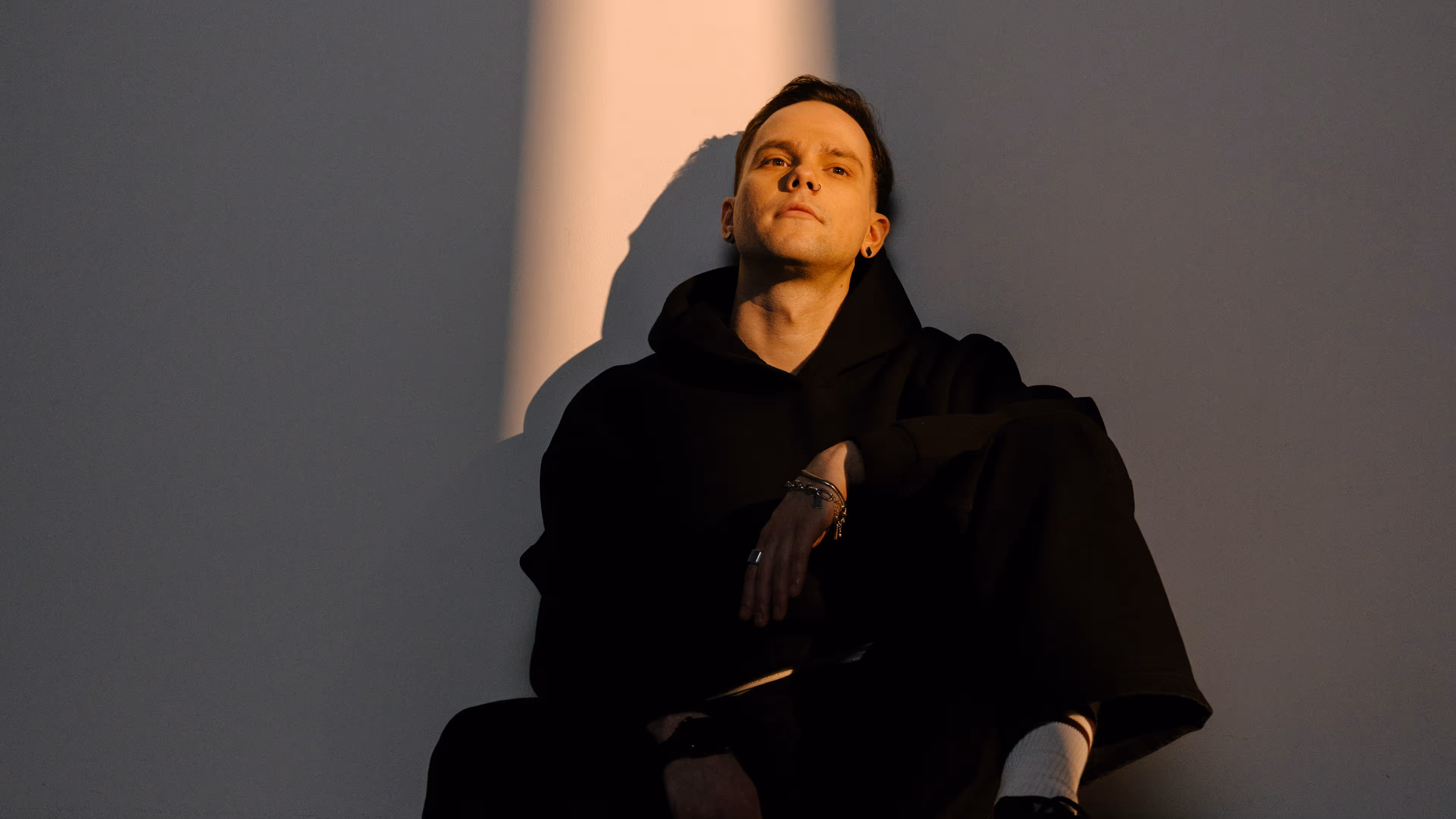 Young man in black hoodie sitting against a wall with shadows cast on his face and body.