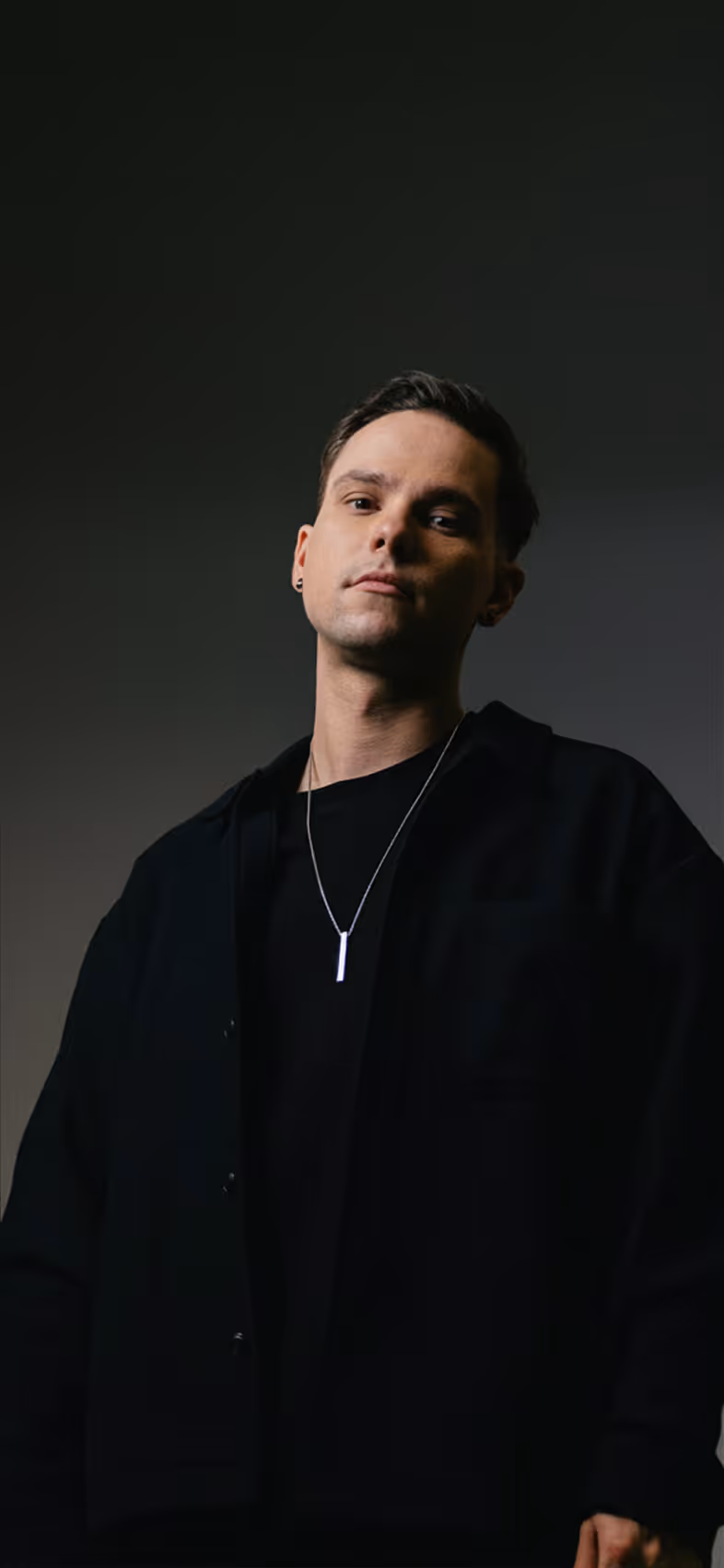 Young man wearing a black shirt and jacket with a silver necklace against a dark background.