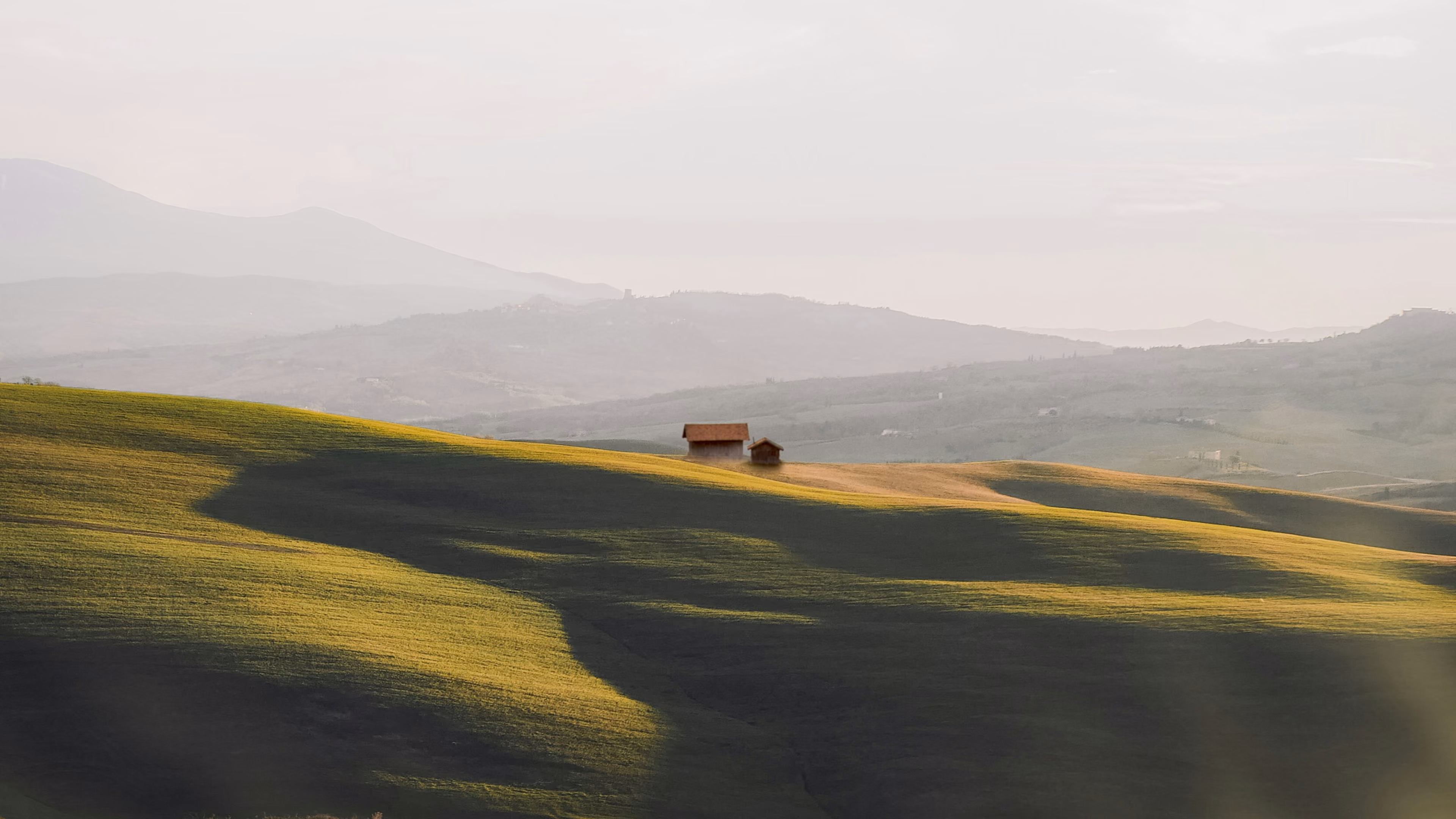 Two small houses on a sunlit green hill with mountains fading in the misty background.