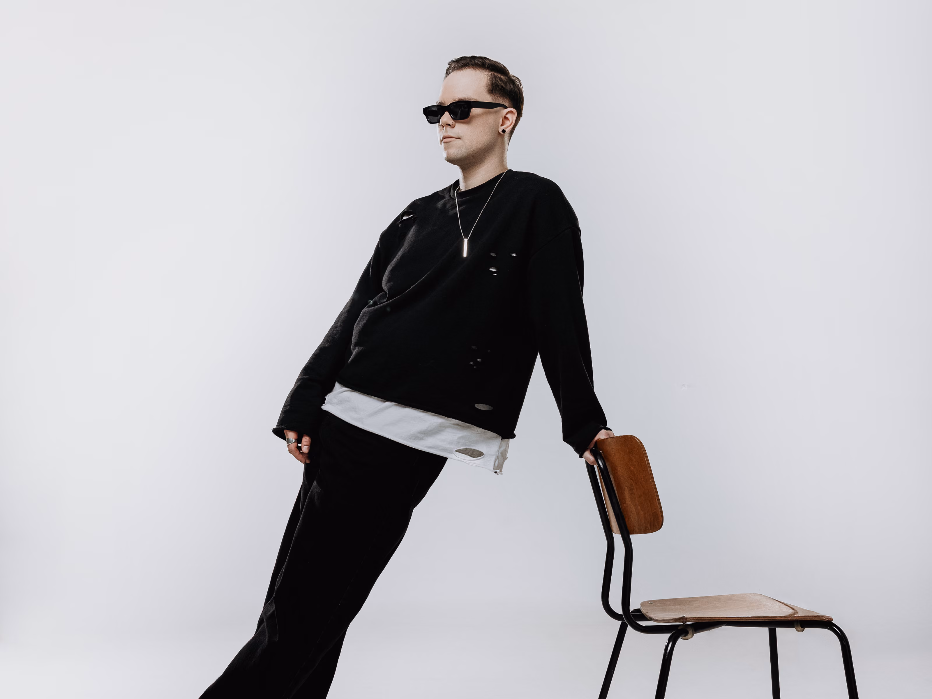 Man in black ripped sweatshirt, sunglasses, leaning backward on a wooden chair against a white background.