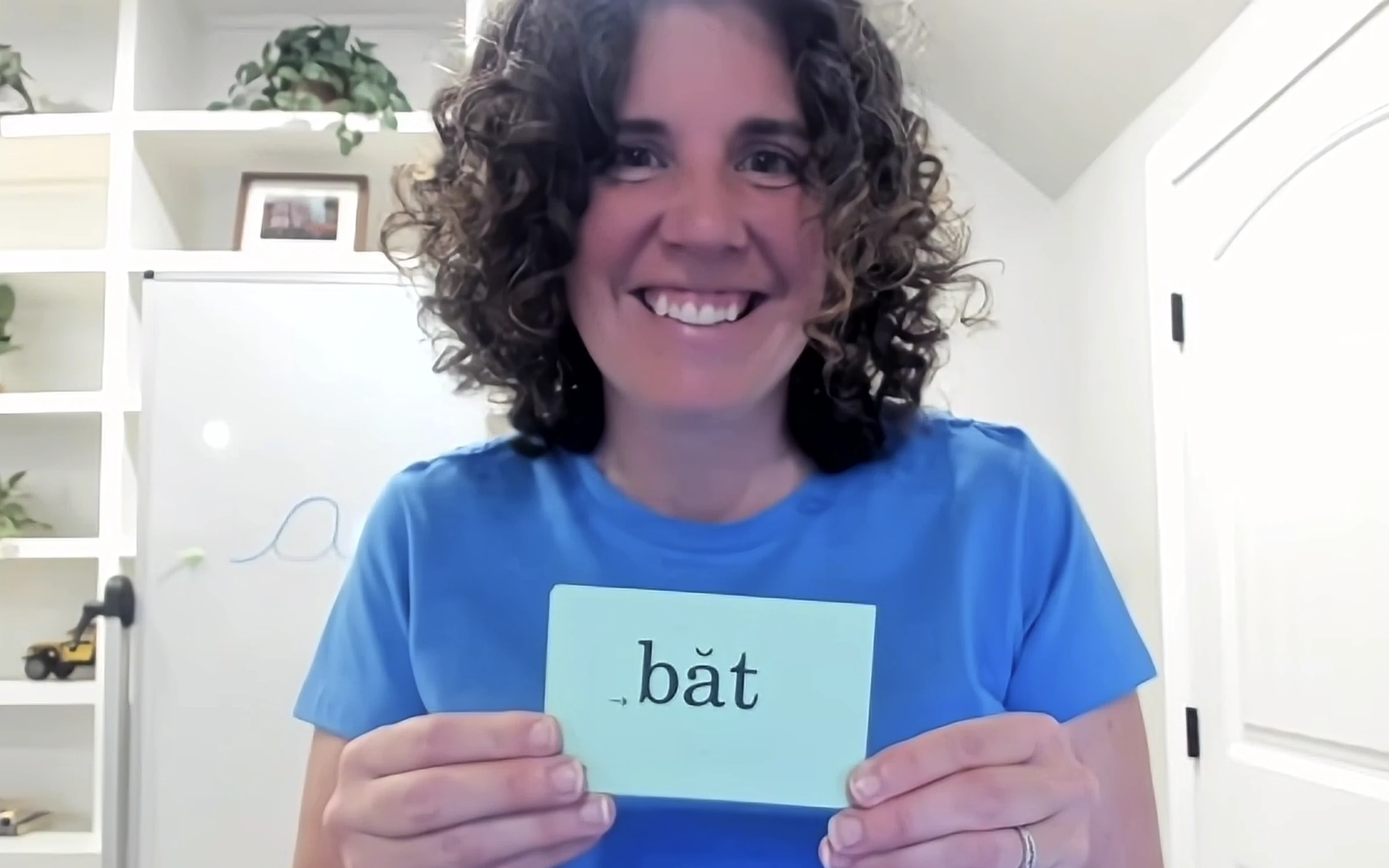 Elizabeth smiling and holding a flashcard reading bat with the short-vowel mark during a virtual session