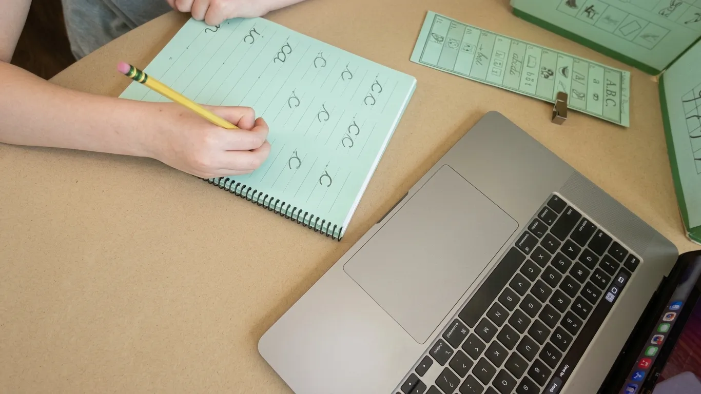 A child's hand writing letter forms in a notebook with a pencil during a virtual session, a sound-card strip beside it and an open laptop at the edge of the table.