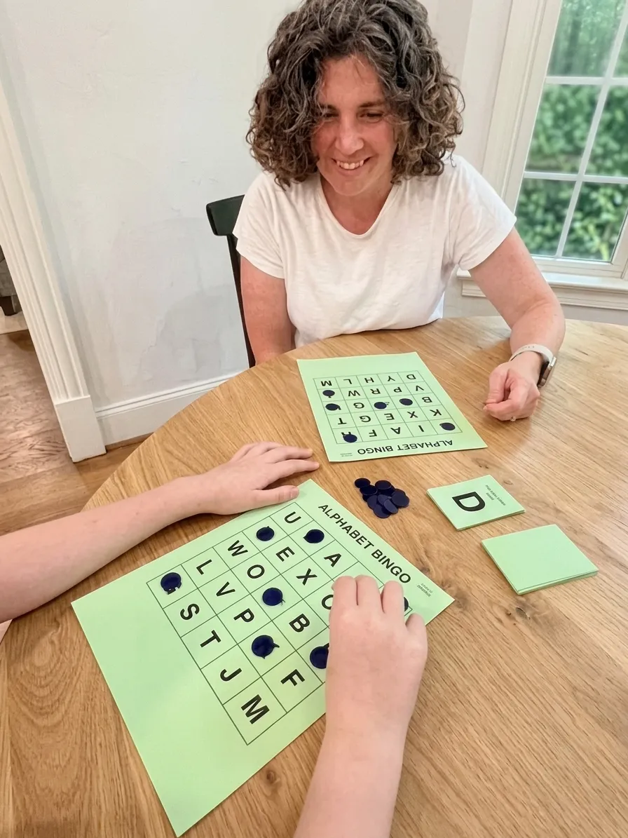Elizabeth across a table from a student, smiling, as the student places counters on an alphabet bingo card.