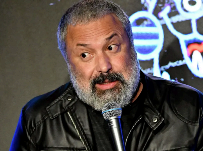 A bearded man in a leather jacket speaking into a microphone on stage