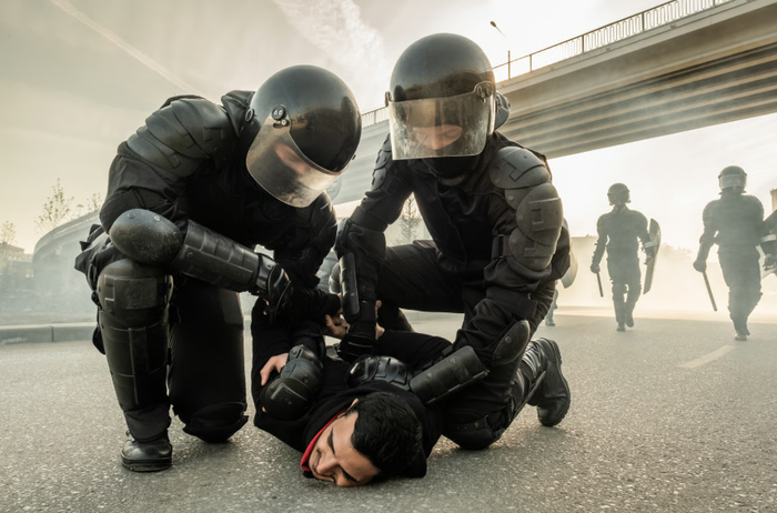 Two police officers in riot gear pinning a man face-down on the pavement