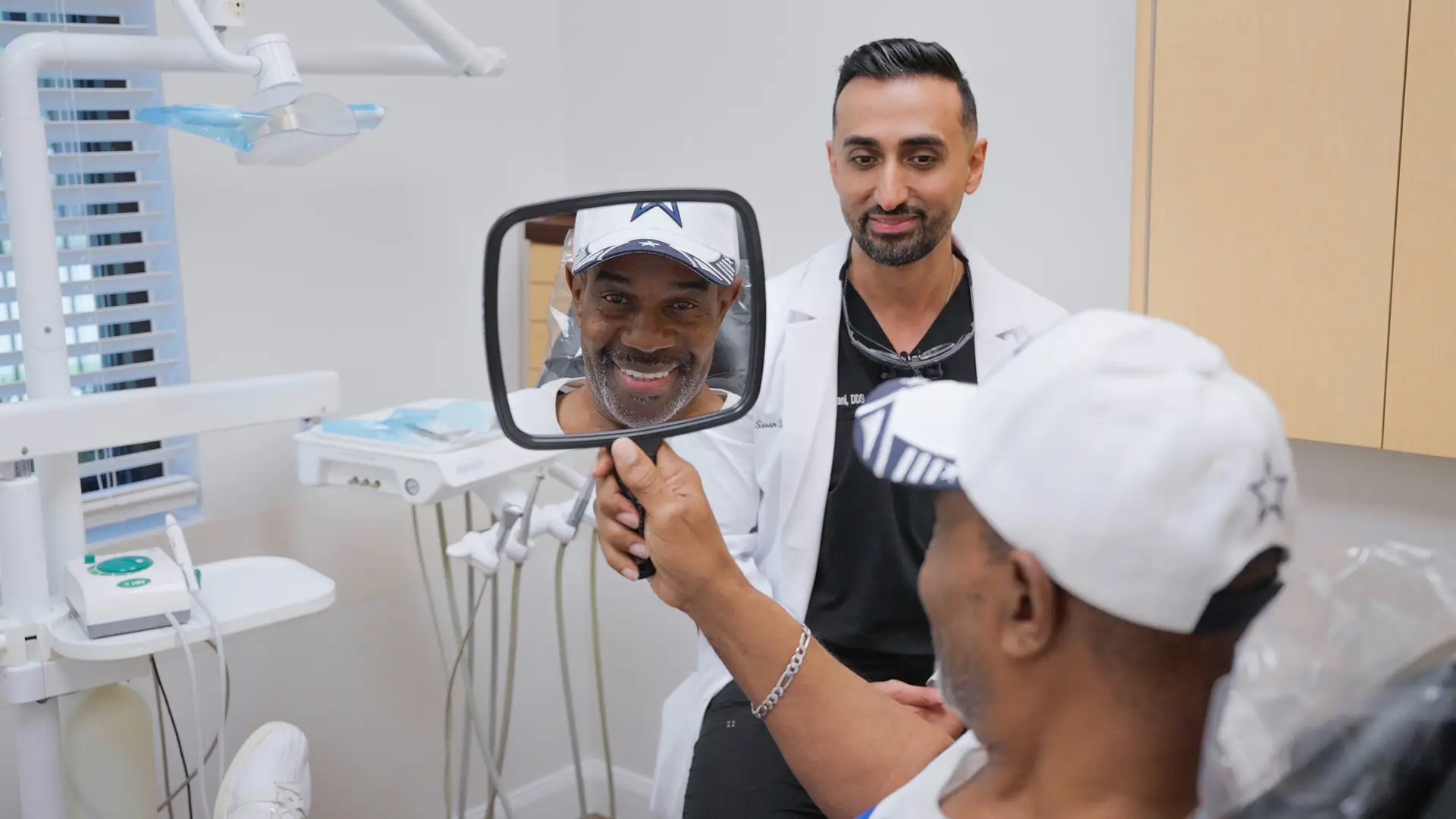 A patient smiles at his own teeth in a hand mirror while Dr. Sasan Bastani looks on
