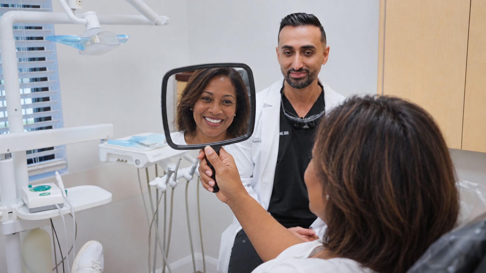 A patient checks his smile in a hand mirror while Dr. Bastani looks on
