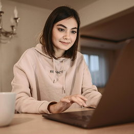 Young woman wearing a beige hoodie working on a laptop in a cozy room.