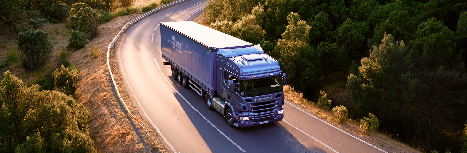 Blue semi-truck driving on a winding road surrounded by green trees and dry grass.