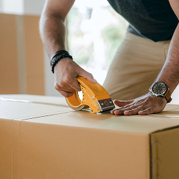 Person sealing a cardboard box with a yellow tape dispenser in a light-filled room.