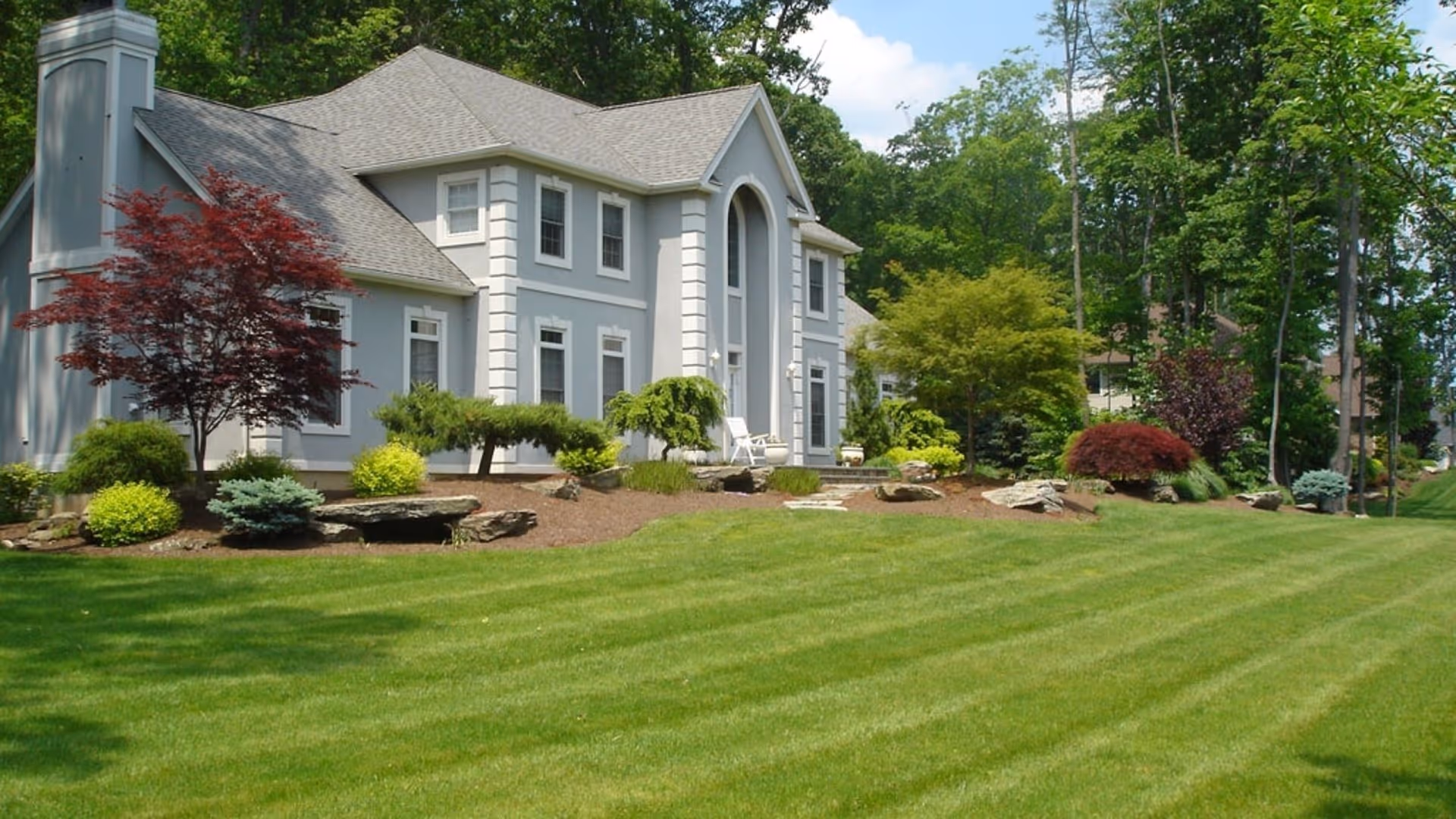 Large gray house with multiple windows, surrounded by manicured lawn, shrubs, and trees in daylight.