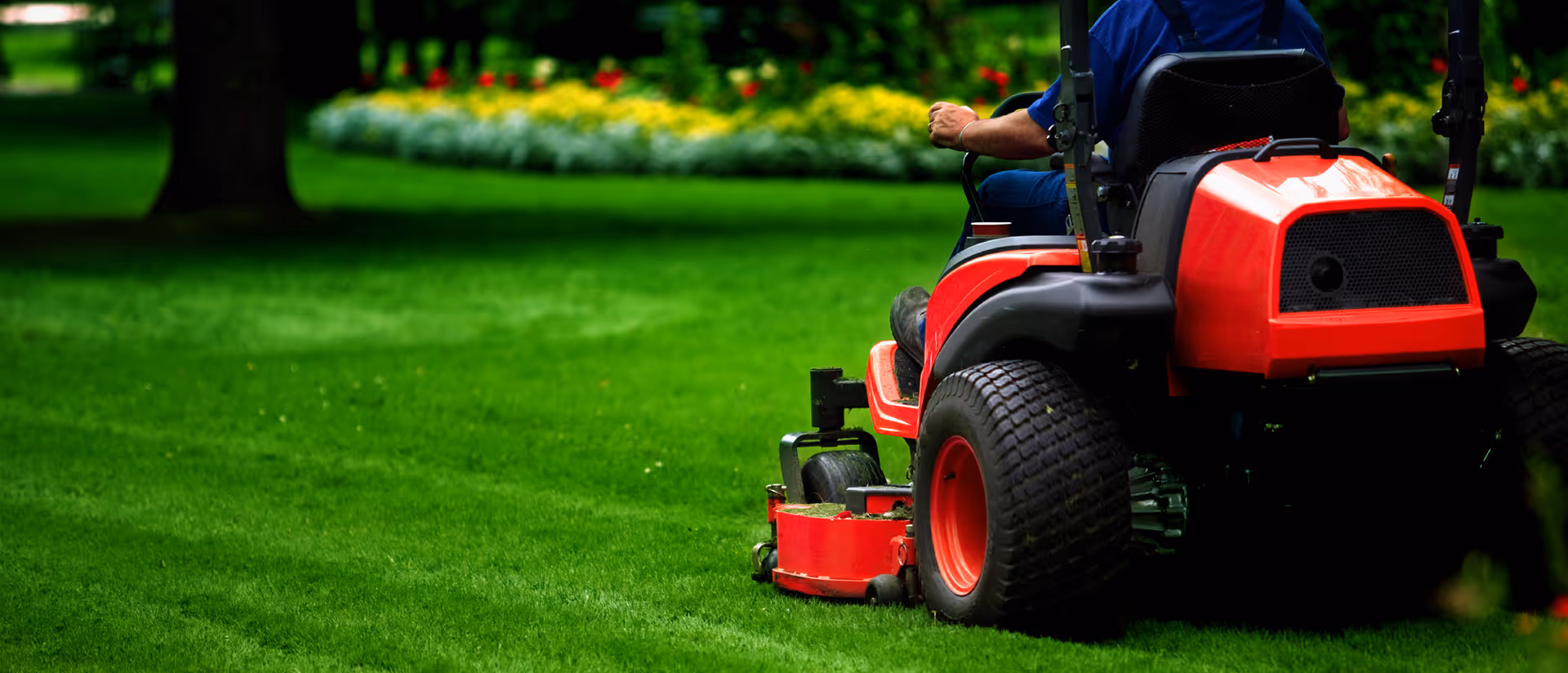 Person riding a red lawn mower cutting bright green grass near colorful flower beds.