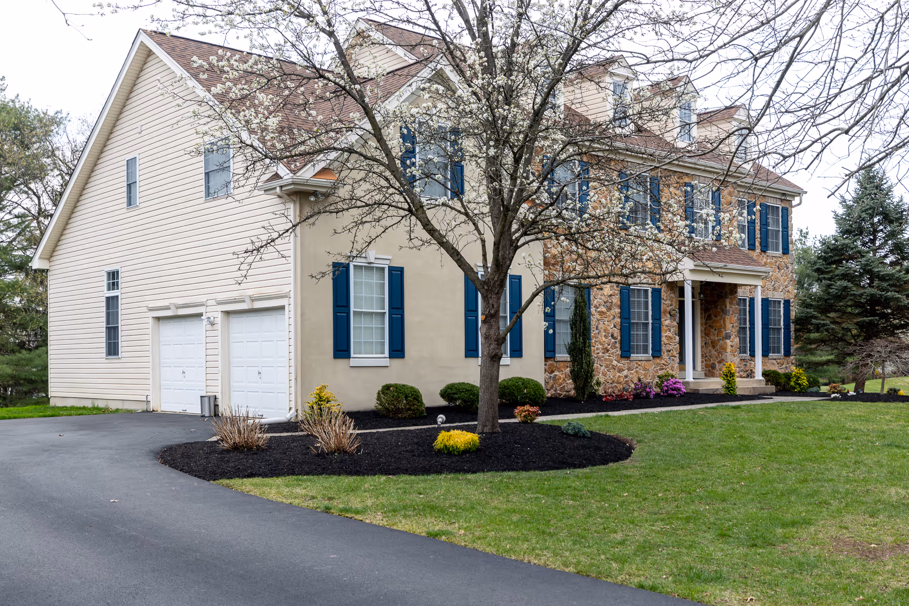 Two-story house with stone and beige siding, blue shutters, a blossoming tree, and a curved driveway.
