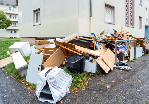 Piled discarded furniture and household items dumped outside a residential building on a wet pavement.