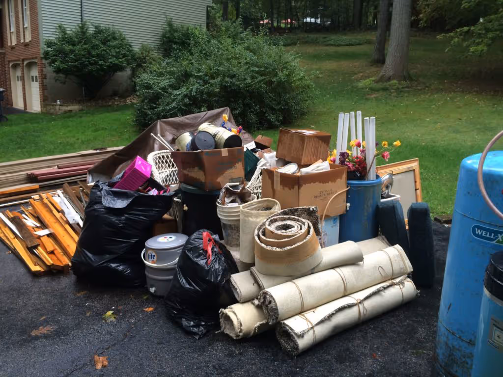 Pile of rolled carpets, black trash bags, boxes, and other junk on a driveway near grass and trees.