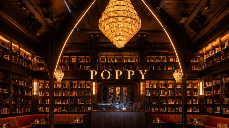 Interior of a dimly lit library or lounge with wooden shelves, chandelier, and a sign reading POPPY.