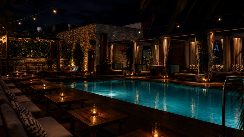 Dimly lit rooftop pool area at night with wooden tables, cushioned seats, and string lights.