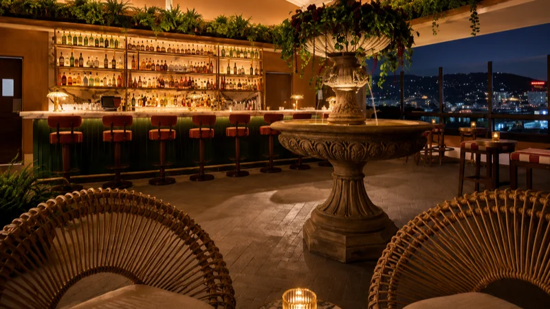 Cozy rooftop bar at night with wicker chairs, central stone fountain, and city lights in the background.