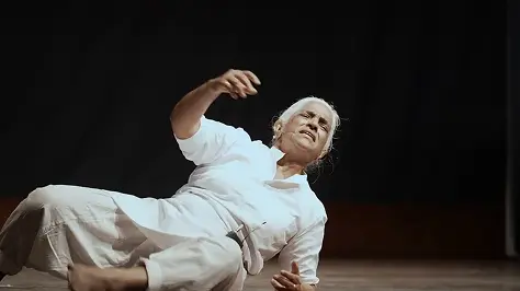 Image of Maya Krishna Rao in a white outfit, mid-performance, expressively gesturing while seated on a dimly lit stage