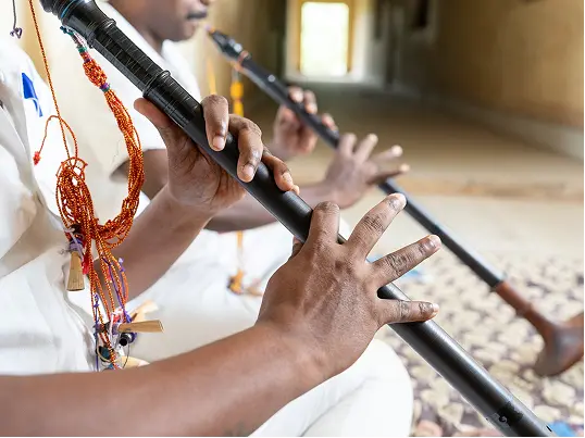 Close-up of two people playing long, black flutes adorned with colorful beads, wearing white clothing.