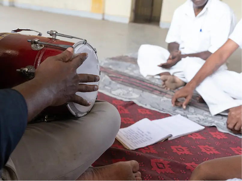 A person plays a hand drum in a casual gathering. Others in white clothing sit cross-legged on patterned mats, with a notebook nearby, suggesting a collaborative musical session.