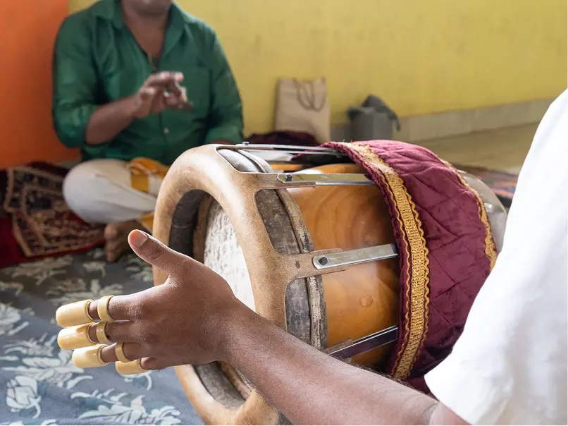Close-up of a musician playing a traditional drum adorned with rings on fingers, with another person in a green shirt sitting cross-legged in the background.