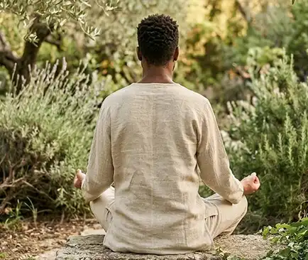 A man in a beige linen top sits in lotus pose on a stone, viewed from behind, meditating in a lush Mediterranean-style garden with herbs and olive trees.