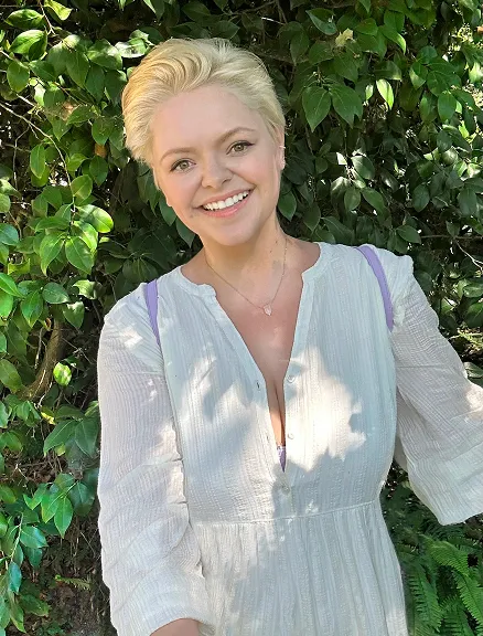 A smiling woman with short blonde hair wearing a white flowy dress stands outdoors in dappled sunlight in front of green leafy foliage, wearing a crystal pendant necklace.
