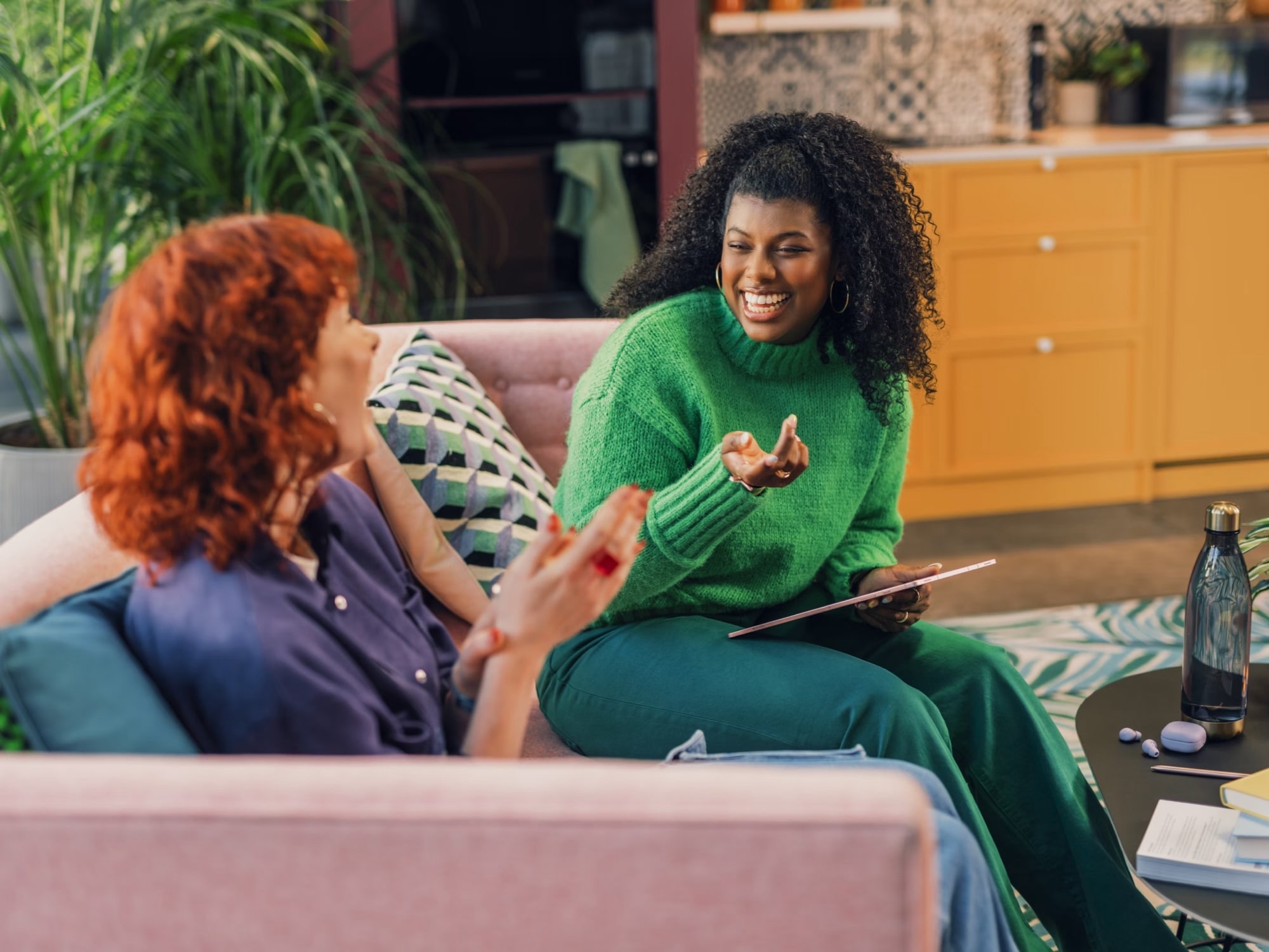 Two women smiling and talking on a pink sofa in a cozy, colorful living room with plants and yellow cabinets.