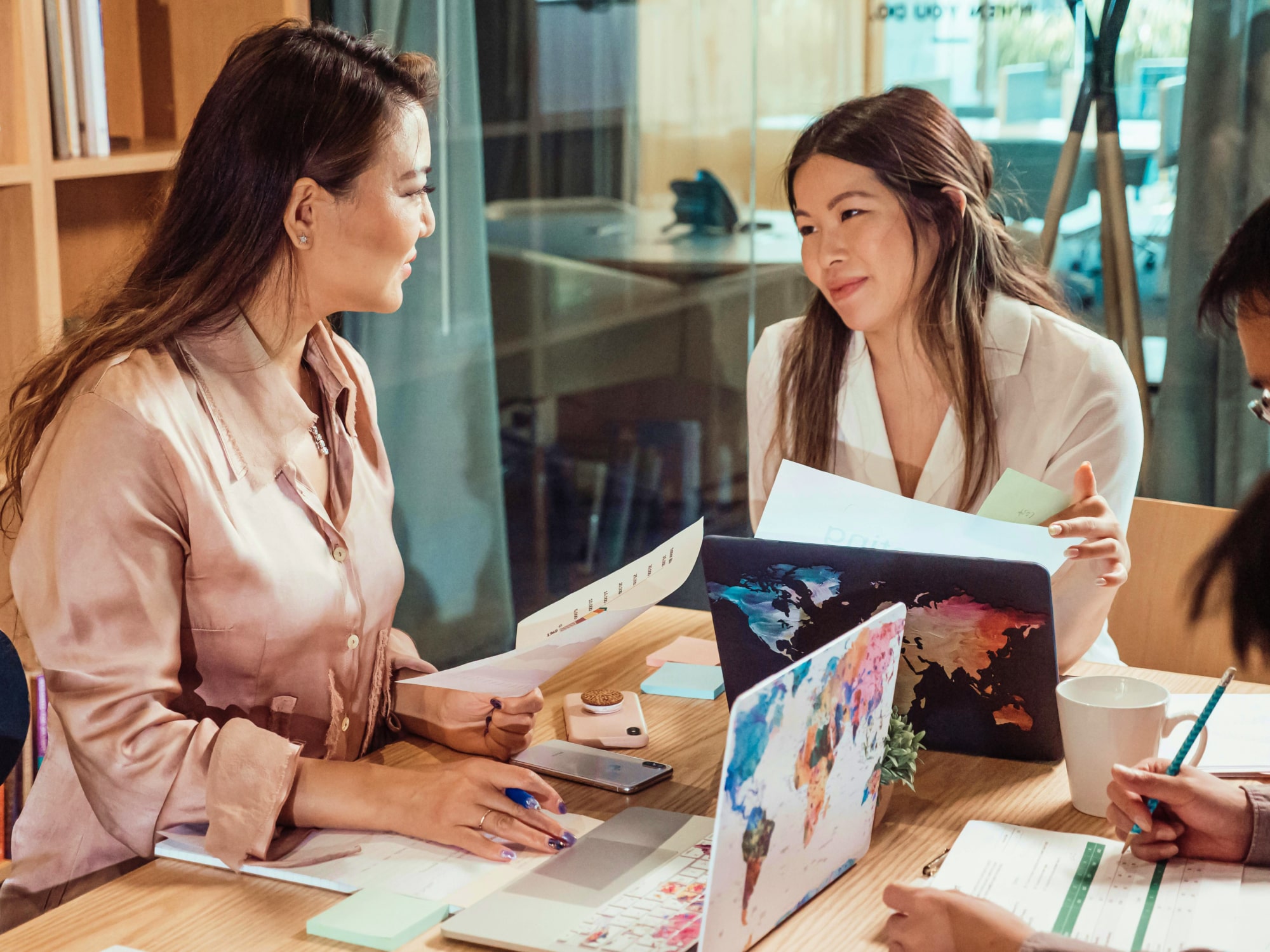Two women discussing documents and laptops with world map covers at a wooden table in an office.